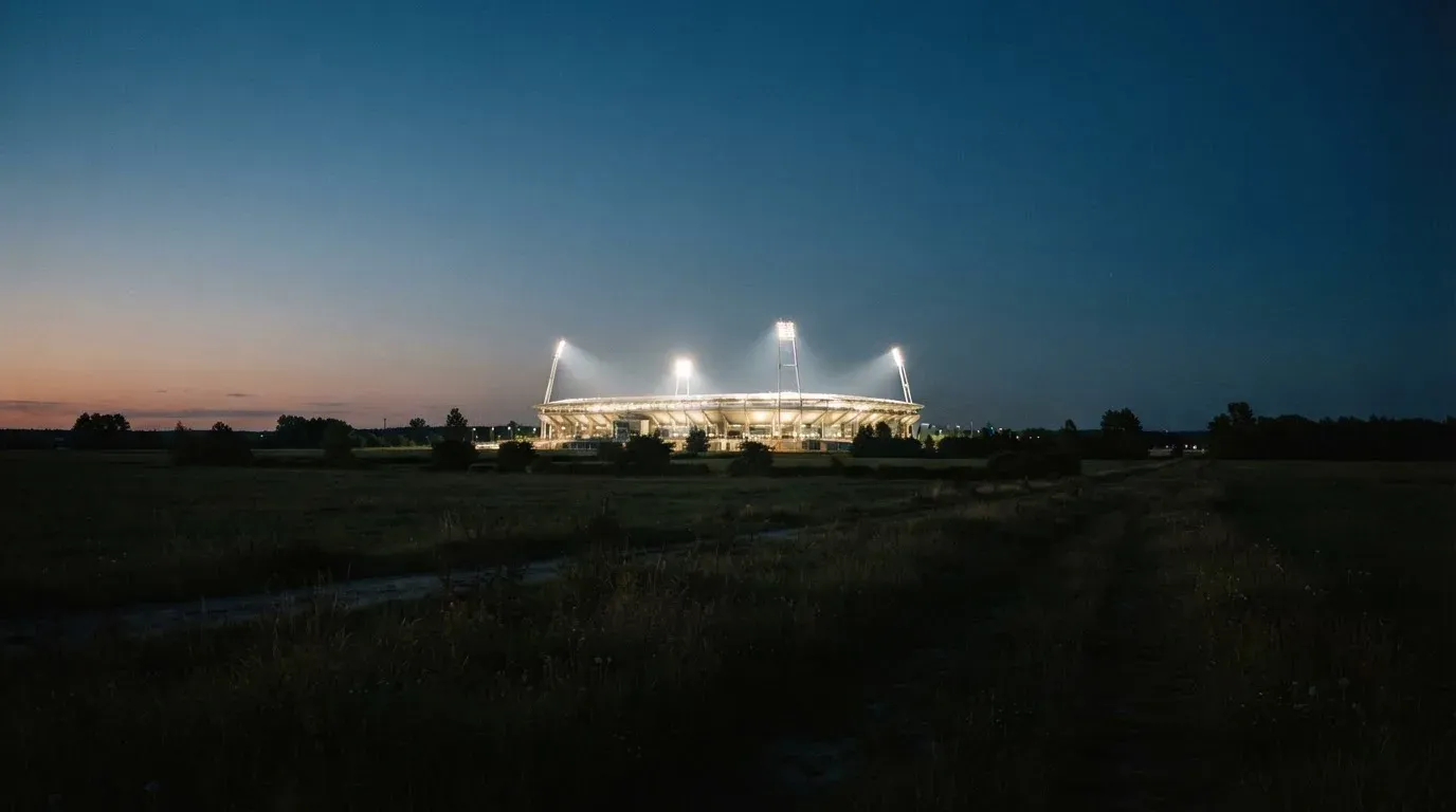 Stadion rozświetlony reflektorami widziany z daleka znad ciemnych pól w pogodny wieczór.