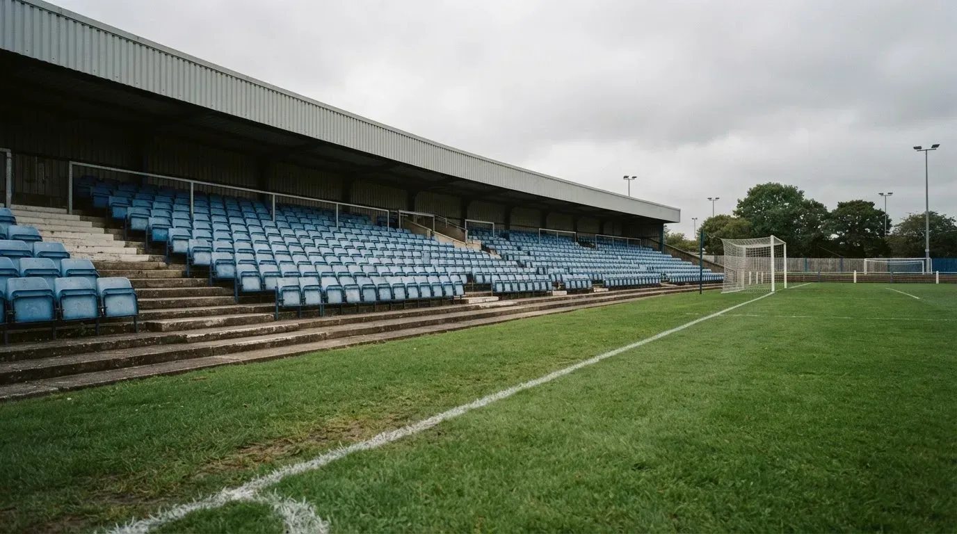 Rzędy pustych plastikowych krzesełek małego stadionu widziane z poziomu murawy pod pochmurnym niebem.