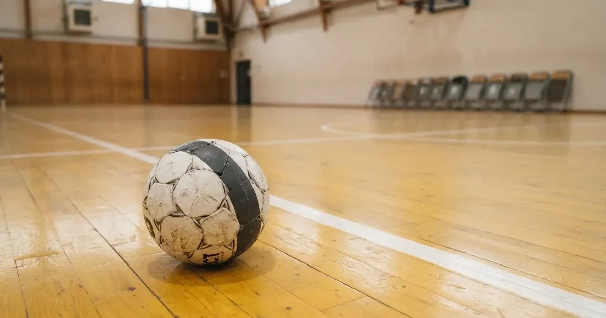 Piłka ręczna leży na żółtym parkiecie sali sportowej, pod ścianą stoją złożone krzesła.