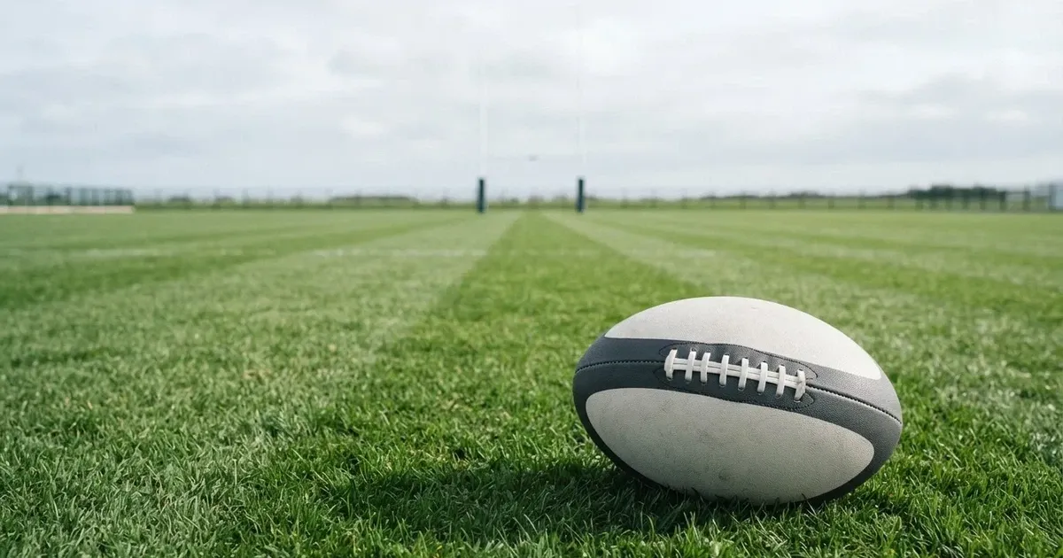 Piłka do rugby leży na pustej murawie, w tle stoją wysokie słupy bramki.