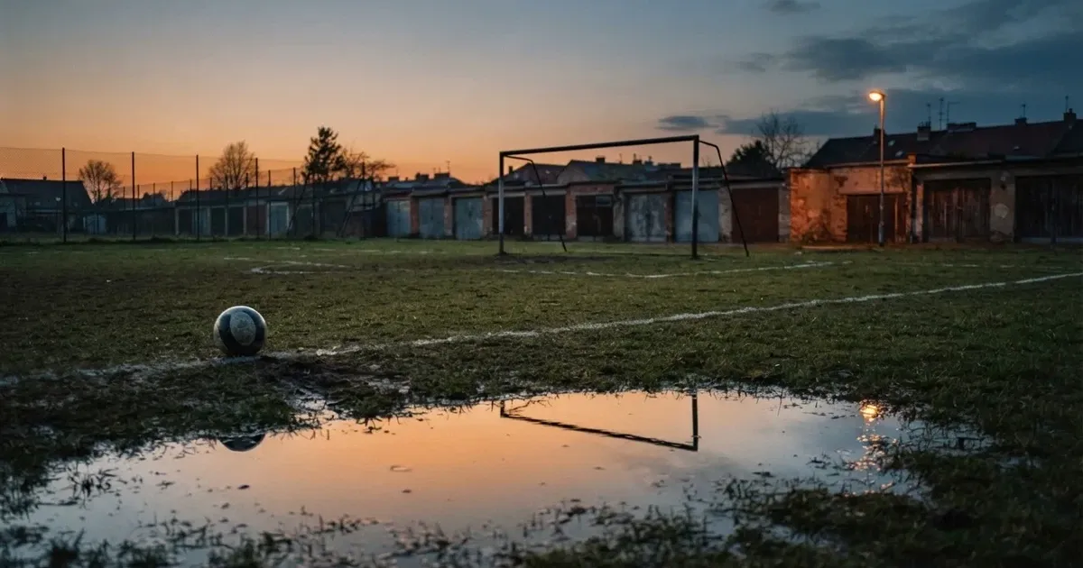 Boisko osiedlowe o zmierzchu, bramka bez siatki, kałuża po deszczu odbija ostatnie światło dnia.