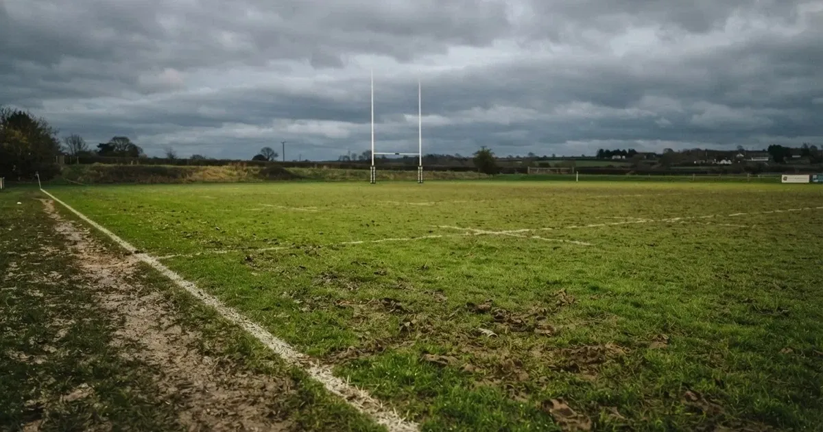 Boisko do rugby ze słupami bramki w oddali pod pochmurnym niebem, mokra trawa i ślady butów.
