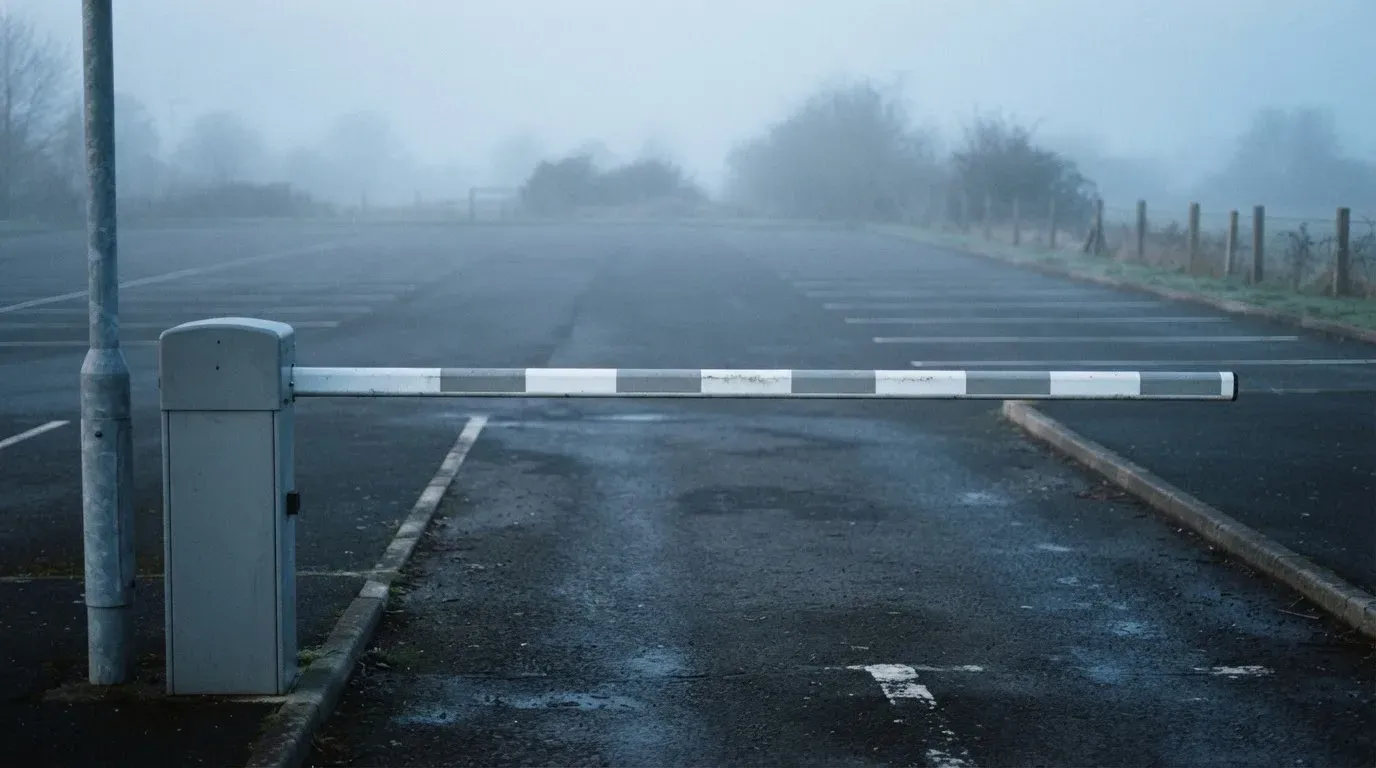 Szlaban w poziomie przy wjeździe na pusty parking o świcie, mgła nad asfaltem.