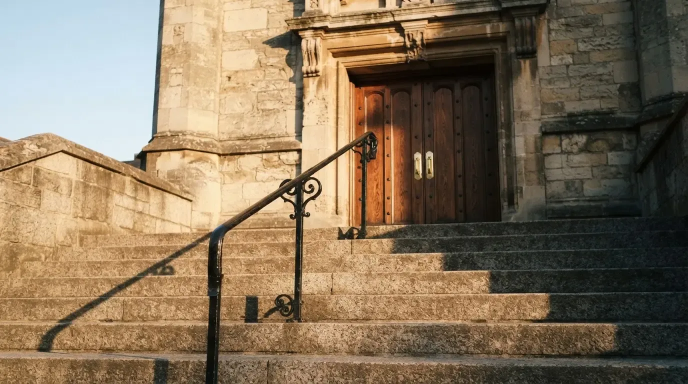 Granitowe schody prowadzące do wysokich drewnianych drzwi budynku w porannym świetle, pusta balustrada.