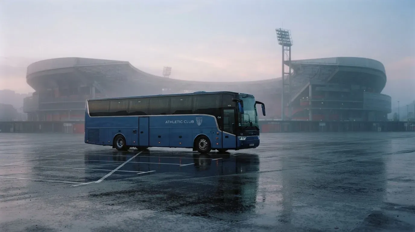 Autokar stoi na parkingu przed stadionem o świcie, rozmyte kontury trybun w tle, pusty plac i mokry asfalt.