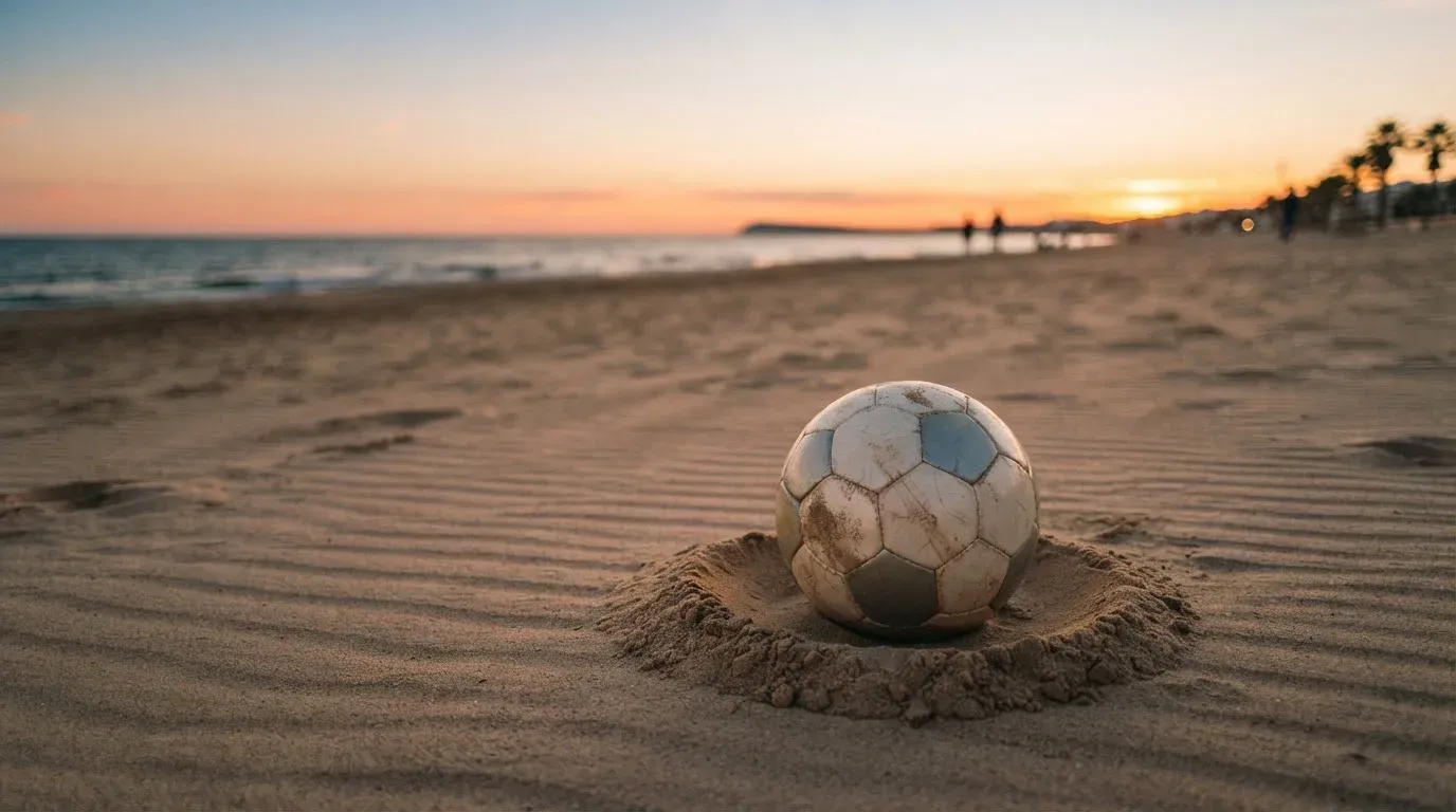 Piłka futbolowa leży na piasku boiska plażowego w ciepłym świetle zachodu, w tle morze i niski horyzont.