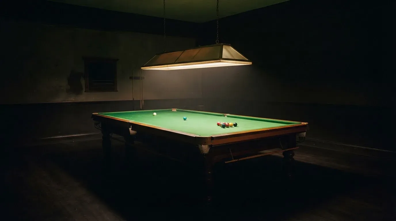 Lampa nad stołem rzuca stożek światła na zielone sukno, reszta pustego pokoju tonie w mroku.